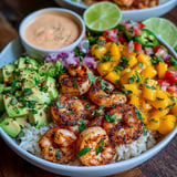 A bowl of food with shrimp, avocado, mango salsa, and lime-chili sauce.